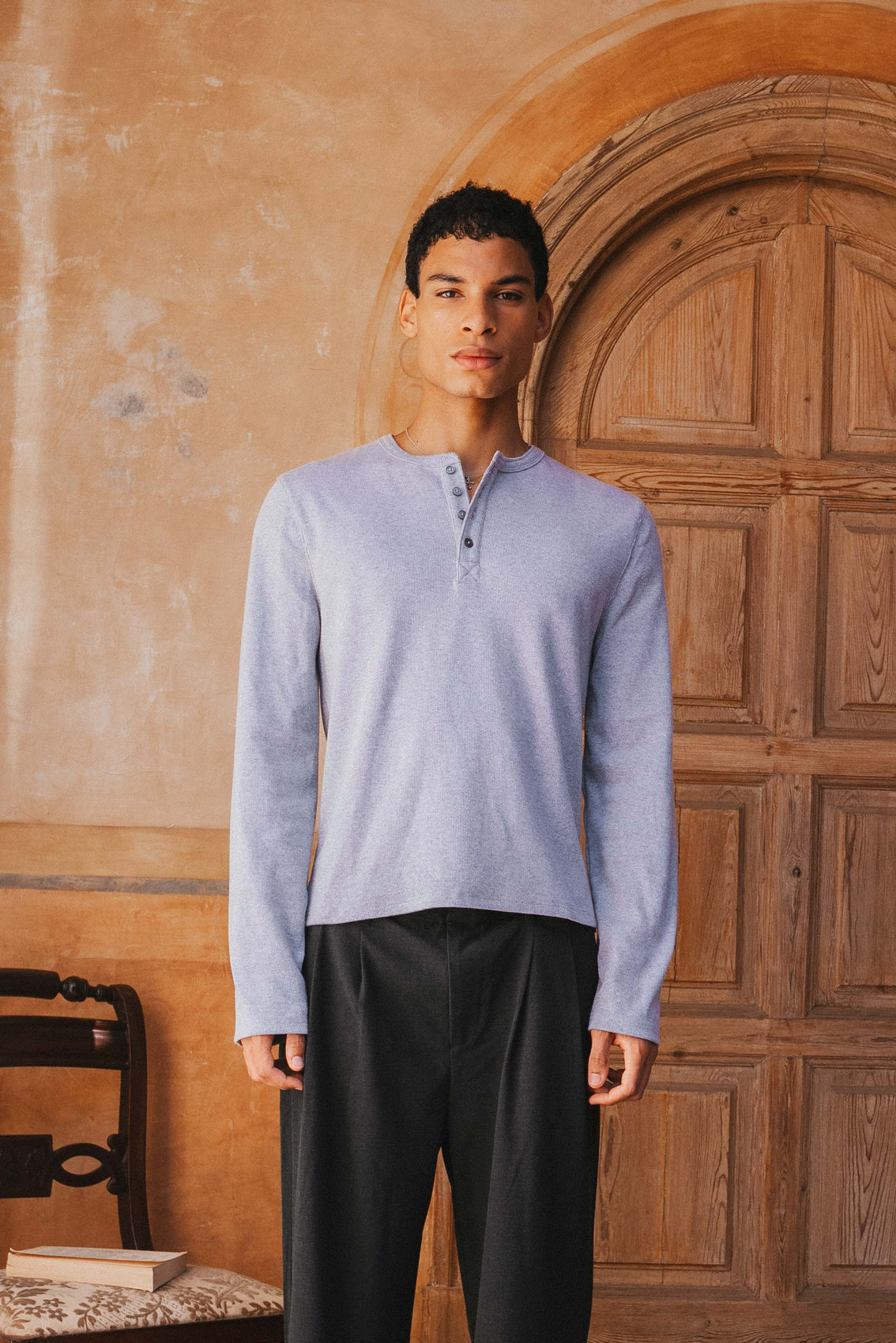 Man wearing a light blue long-sleeve shirt and dark pants standing in front of a wooden door.