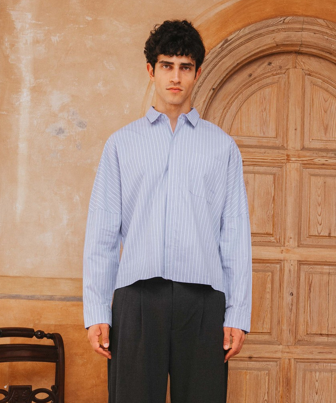 Man wearing a light blue striped shirt and dark pants standing in front of a wooden door.