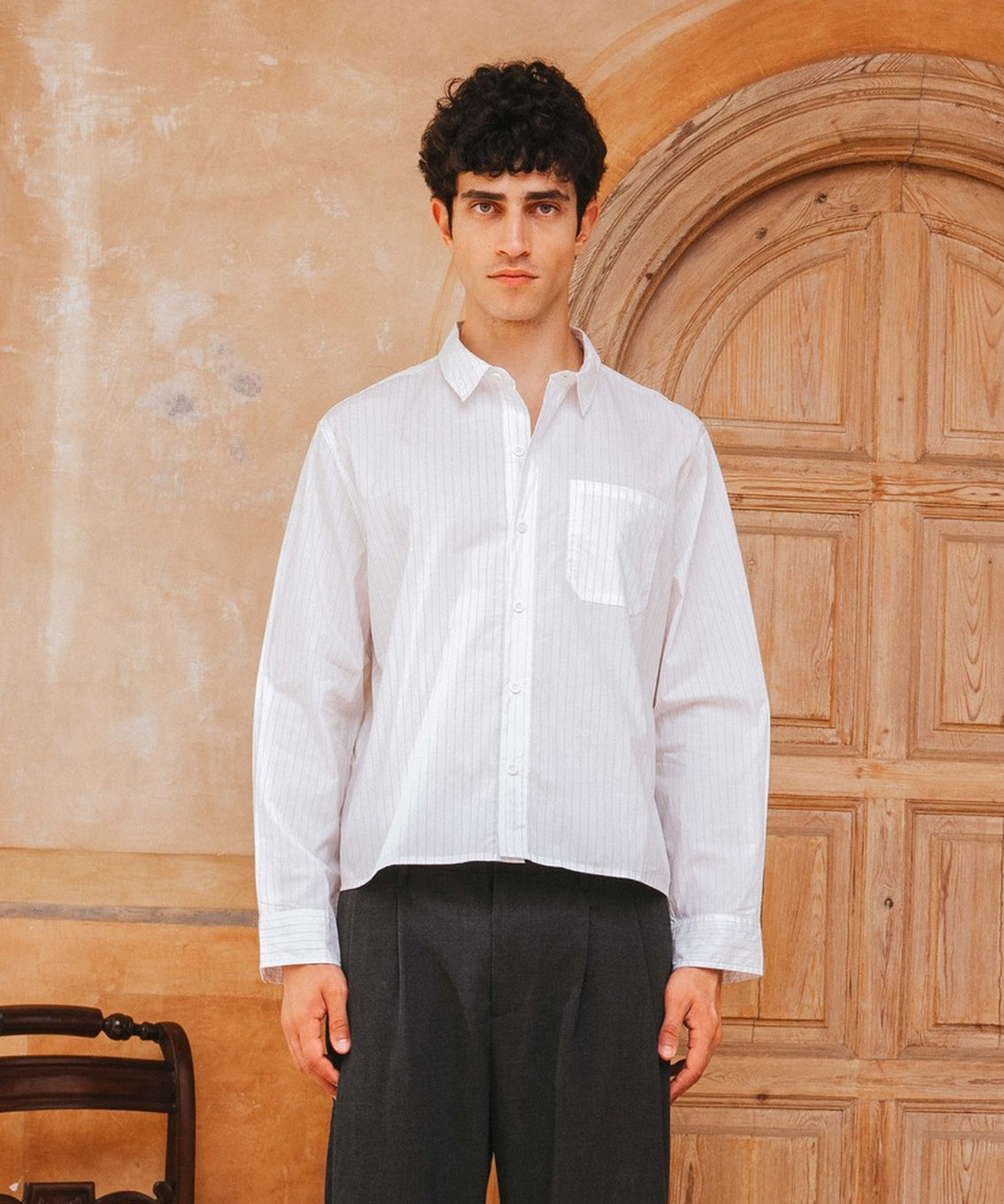 Man wearing a white shirt and black pants standing in front of a wooden door.