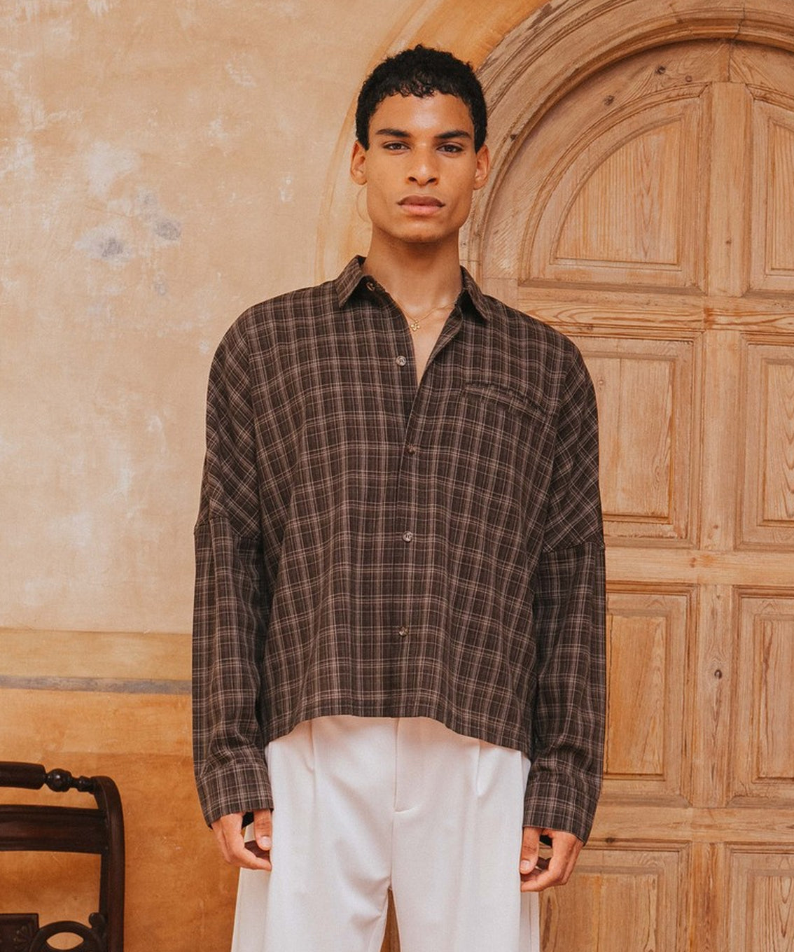 Man wearing a plaid shirt and white pants standing in front of a wooden door.