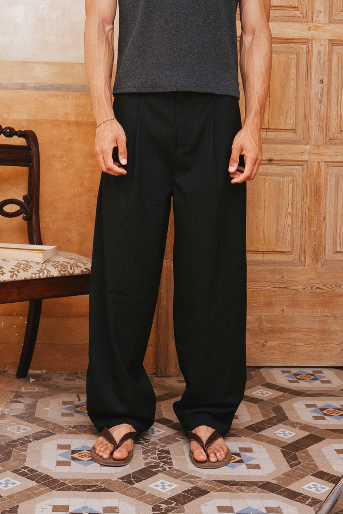 Person wearing black pants and a gray tank top standing in a room with wooden paneling and patterned floor.